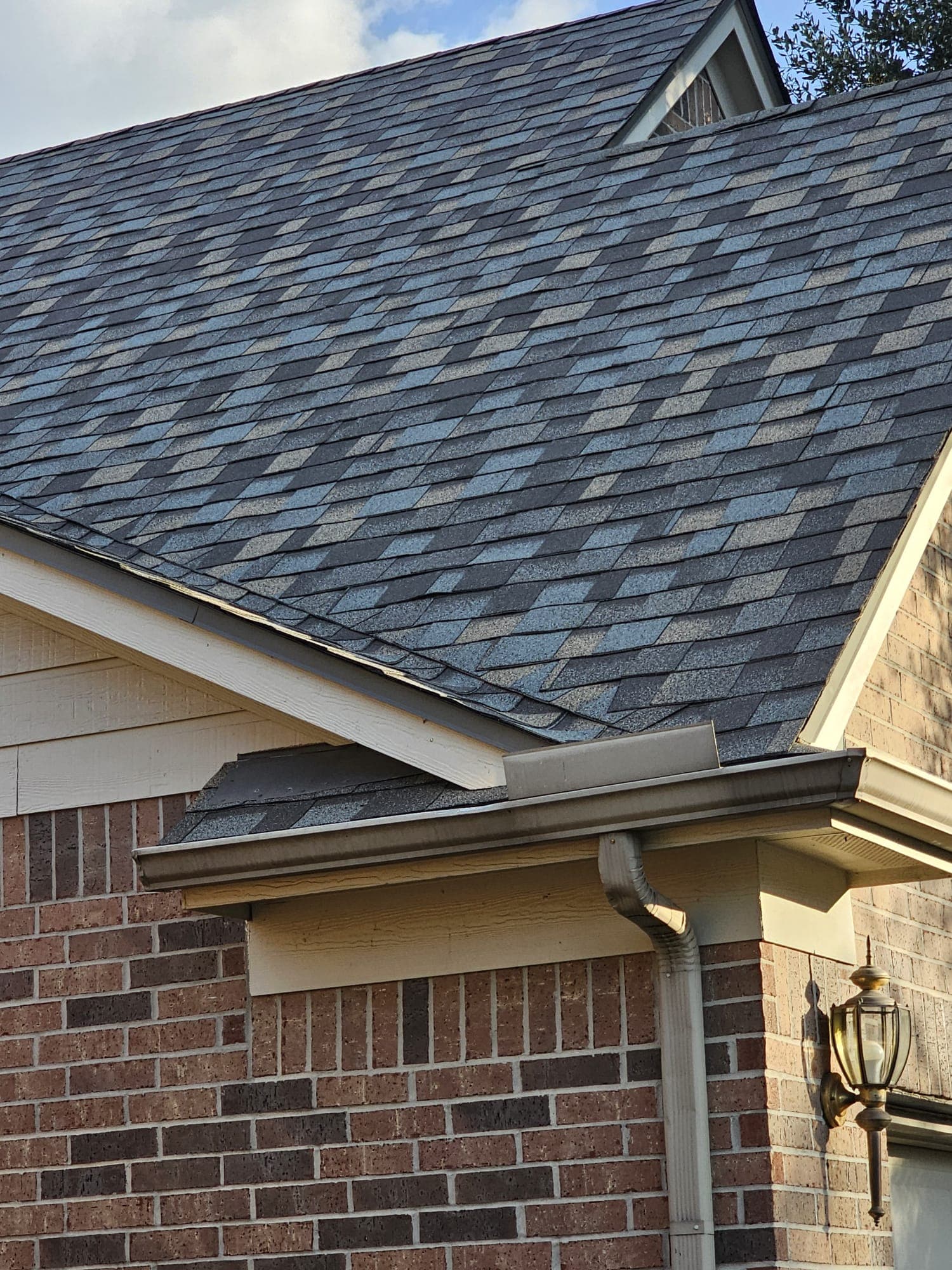 Close-up detail of new dark-grey architectural shingles meeting a brown gutter and coach lamp on a brick home